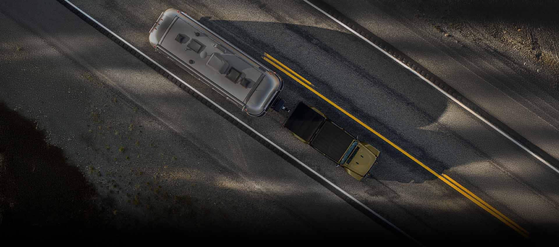 An overhead angle of a olive green 2026 Jeep Gladiator traveling down a highway, towing a large travel trailer.