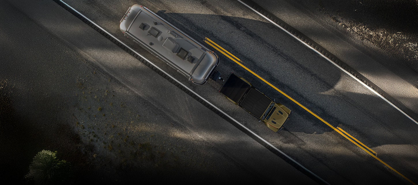 An overhead angle of a olive green 2026 Jeep Gladiator traveling down a highway, towing a large travel trailer.
