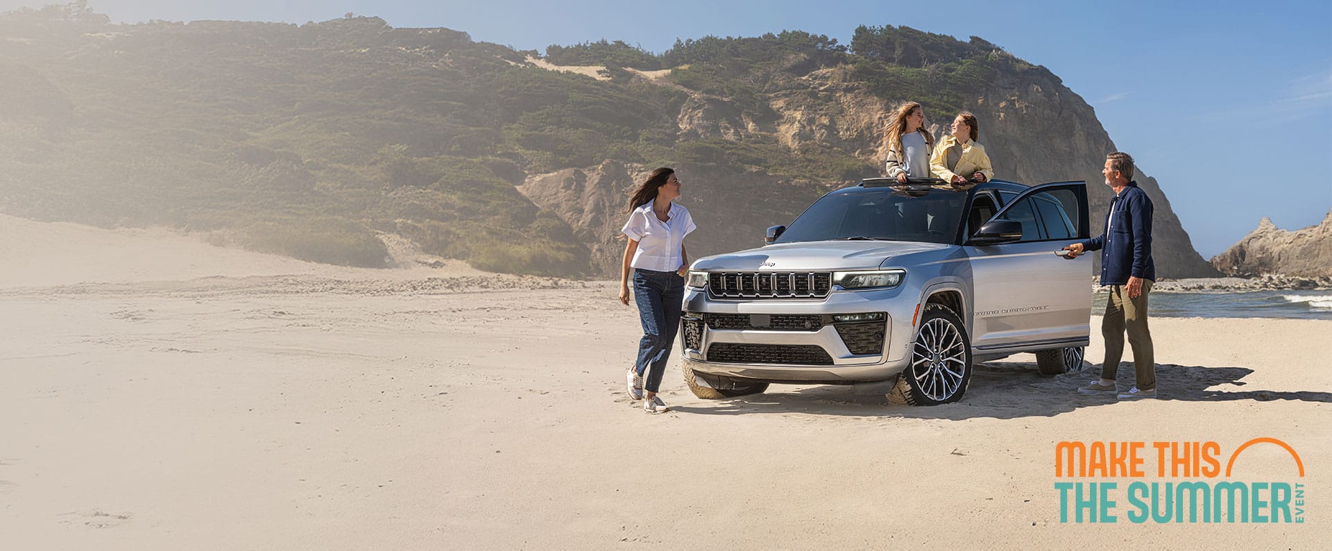 A silver 2026 Jeep Grand Cherokee L Summit, parked on a sandy beach with a couple outside the SUV and two young girls standing up inside the vehicle, through the open sunroof. Make This The Summer Event.