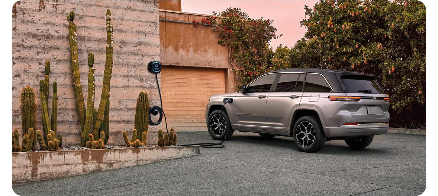 A silver 2026 Jeep Grand Cherokee Summit 4xe parked in the driveway of a large home with the charging cord from a wall-mounted charging station plugged into the vehicle's charging port.
