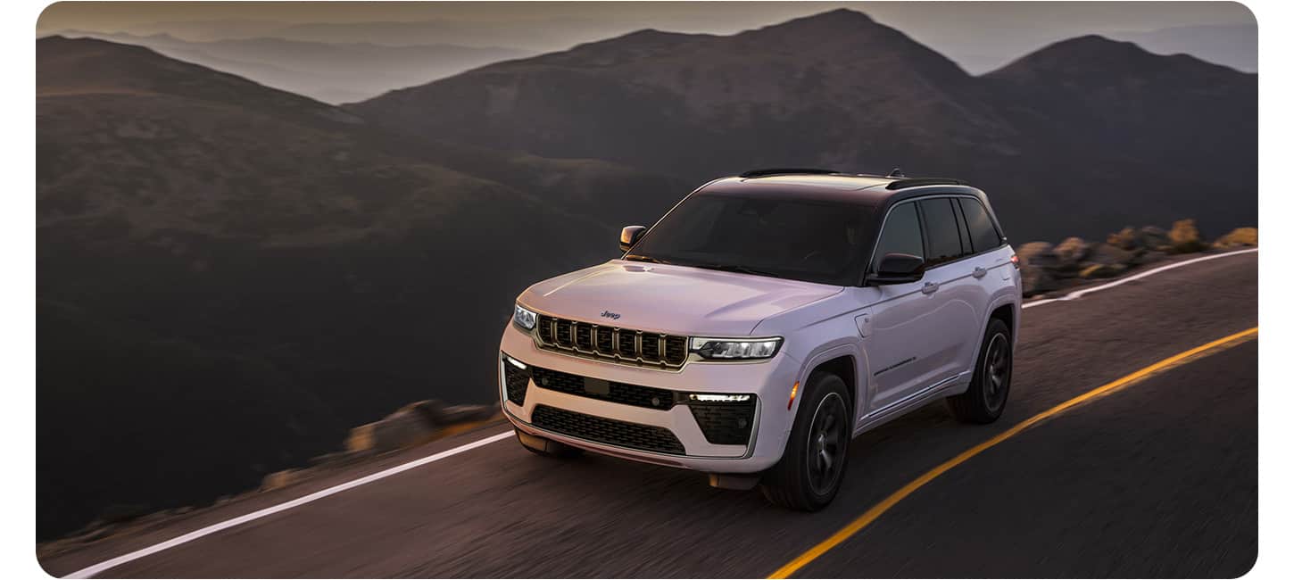 A white 2026 Jeep Grand Cherokee Summit 4xe traveling down a mountain road at dusk.