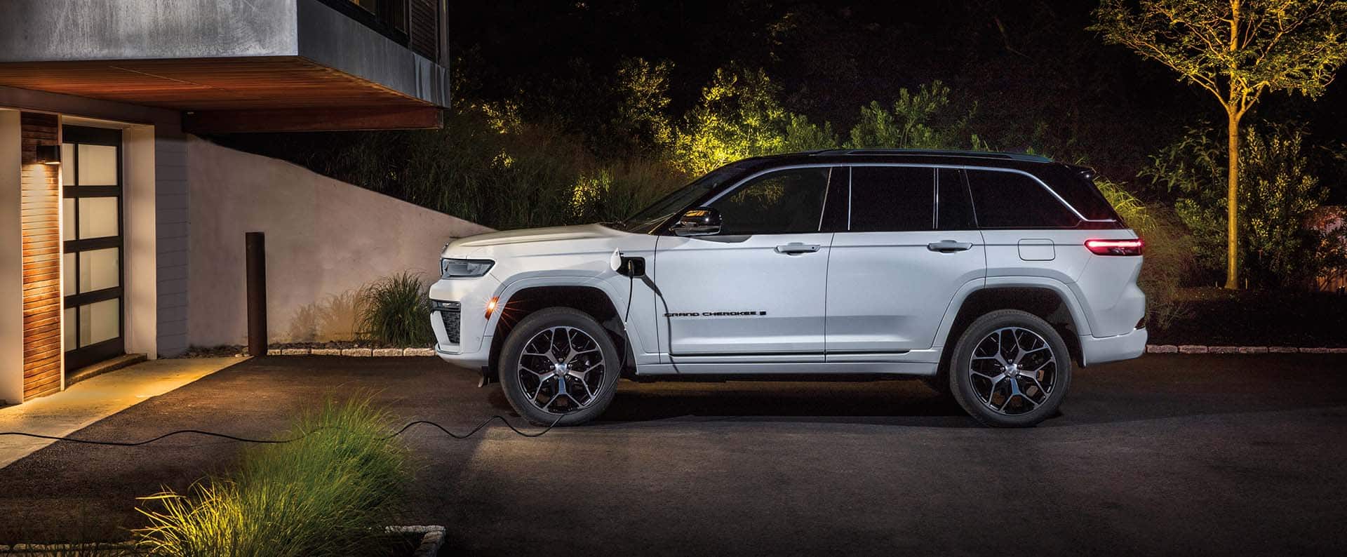 A driver-side profile of a white 2026 Jeep Grand Cherokee Summit 4xe parked in the driveway of a large home with a charging cord plugged into the vehicle's charging port.