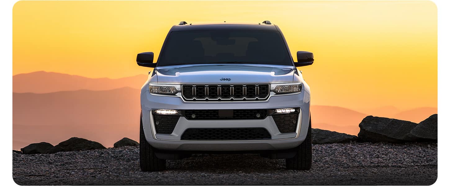 A head-on angle of a silver 2026 Jeep Grand Cherokee Summit on a trail off-road, with two geese blocking its path and a duck on its roof.