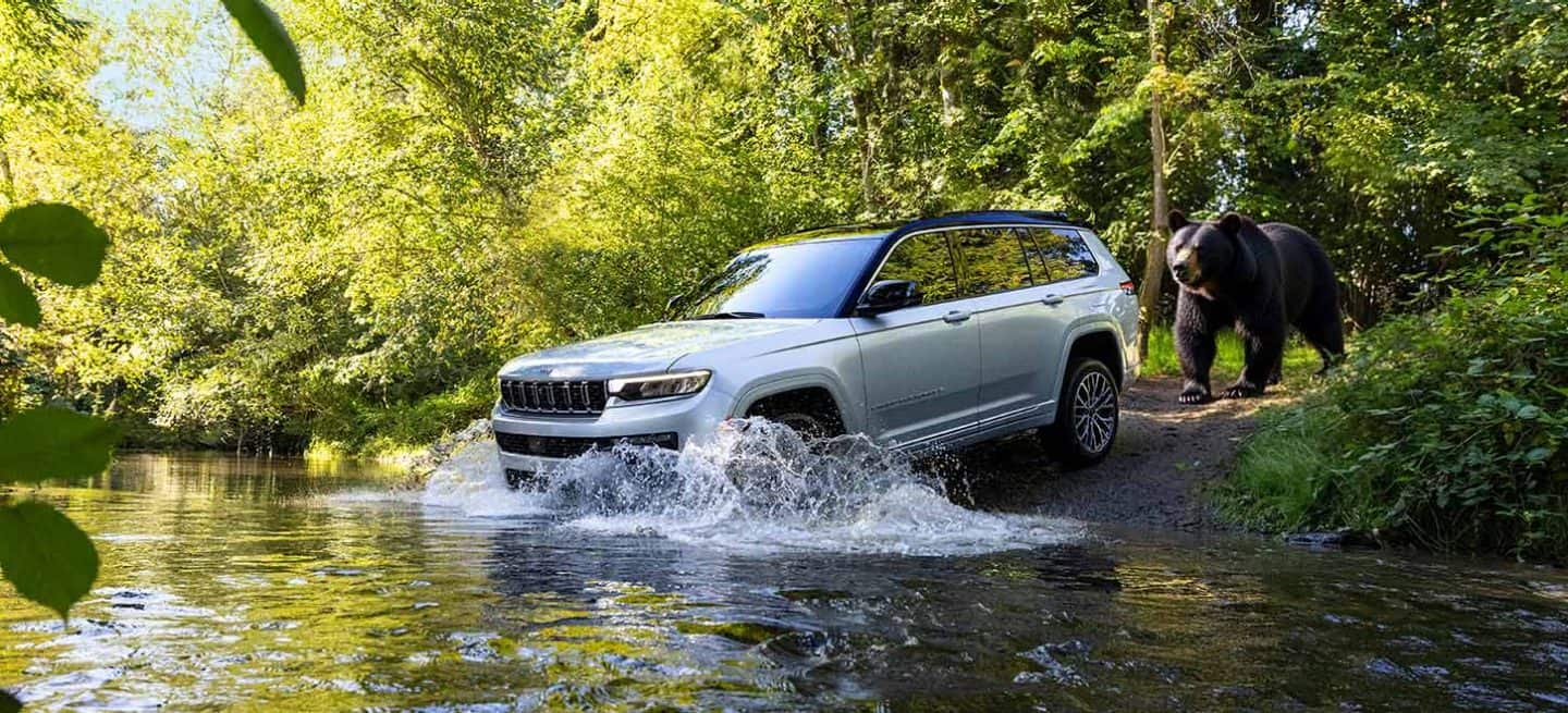 Display A silver 2026 Jeep Grand Cherokee Summit entering a stream, with a large black bear right behind it.