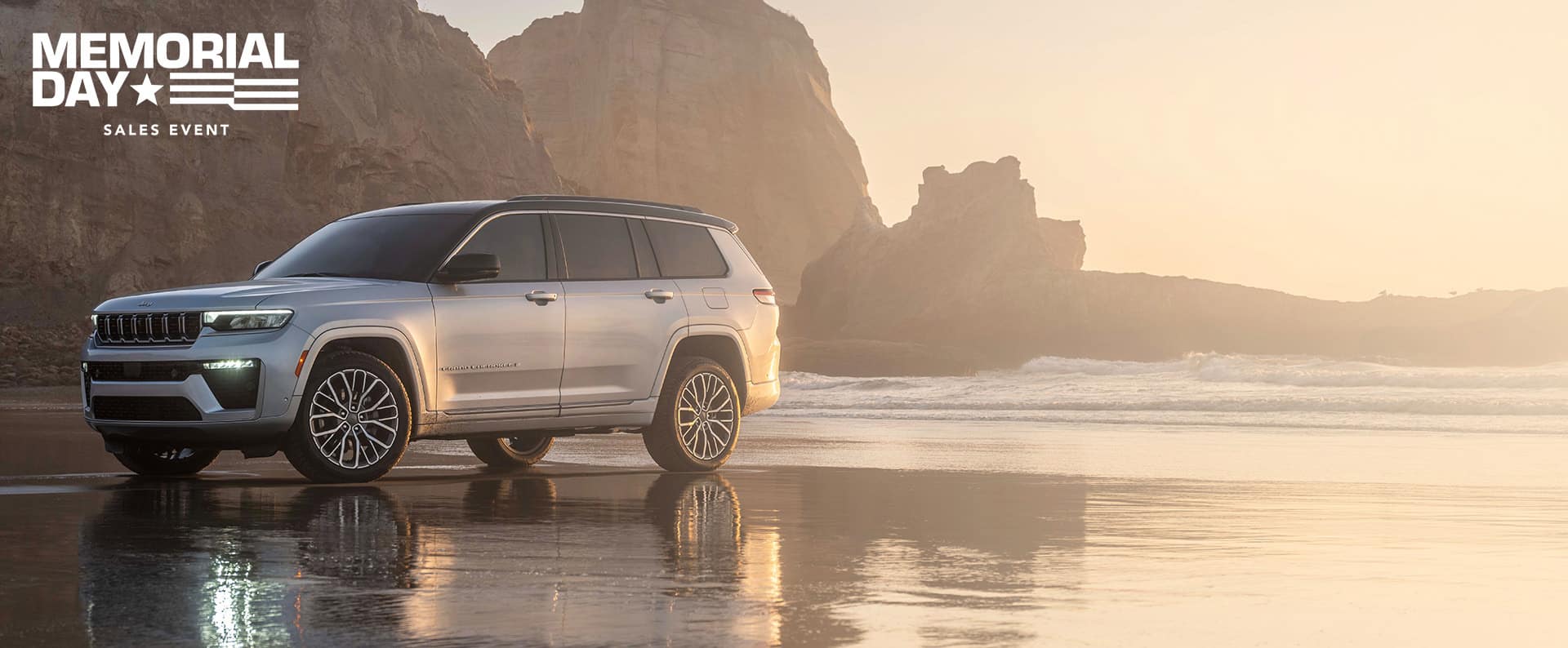 A silver 2026 Jeep Grand Cherokee Summit, parked on a wet beach with rock formations in the background. The Memorial Day Sales Event.