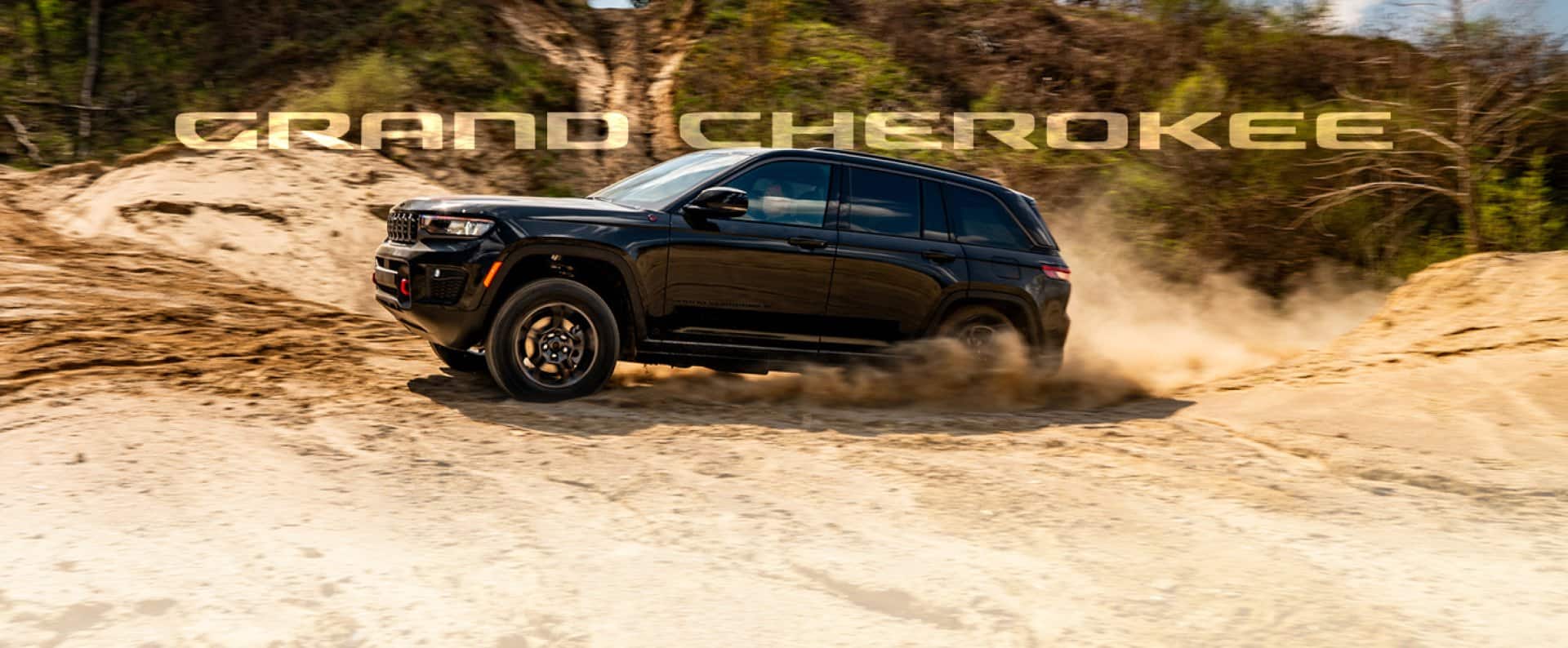 A black Jeep Grand Cherokee Trailhawk kicks up dust as it drives along a dirt road. The words 'GRAND CHEROKEE' are superimposed behind it.