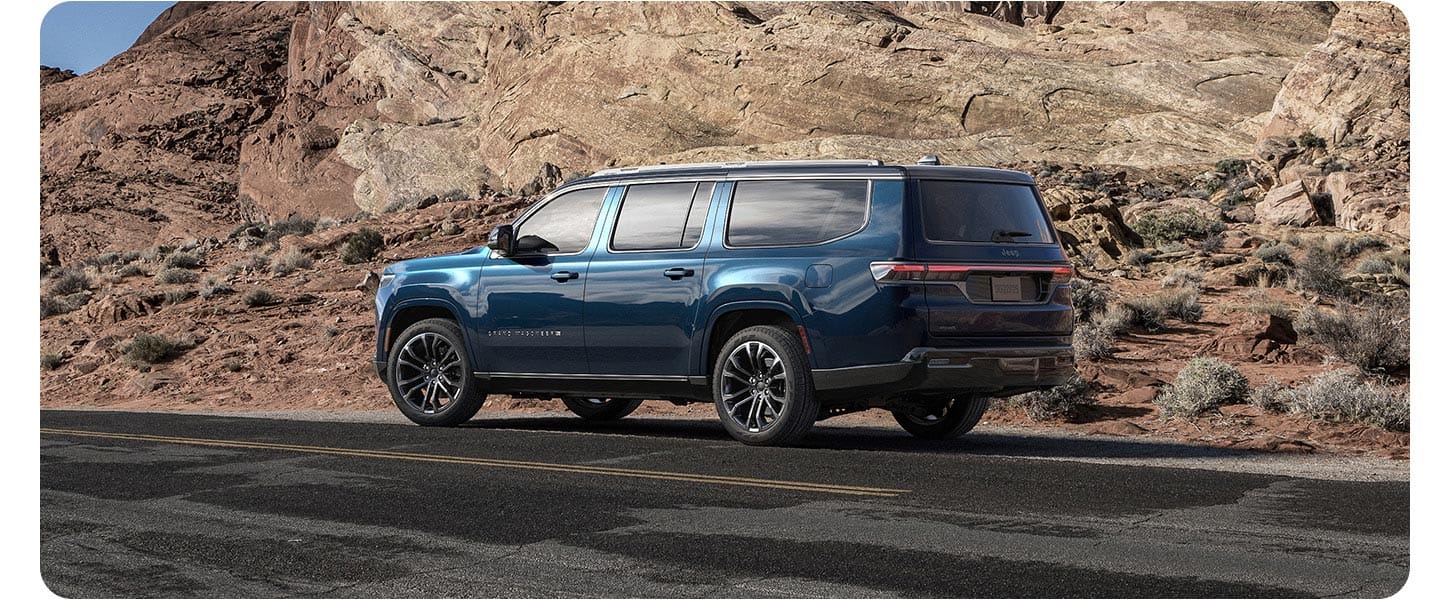 An angled driver-side profile of a blue 2026 Jeep Grand Wagoneer Summit Reserve Obsidian parked on the shoulder of a highway in the mountains.