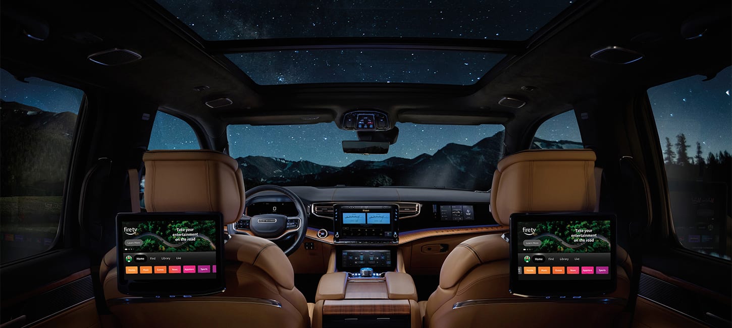 Wide interior view of a Grand Wagoneer Summit Reserve from the rear seat at night, with tan leather-trimmed seats, steering wheel, a panoramic sunroof, and multiple infotainment screens.