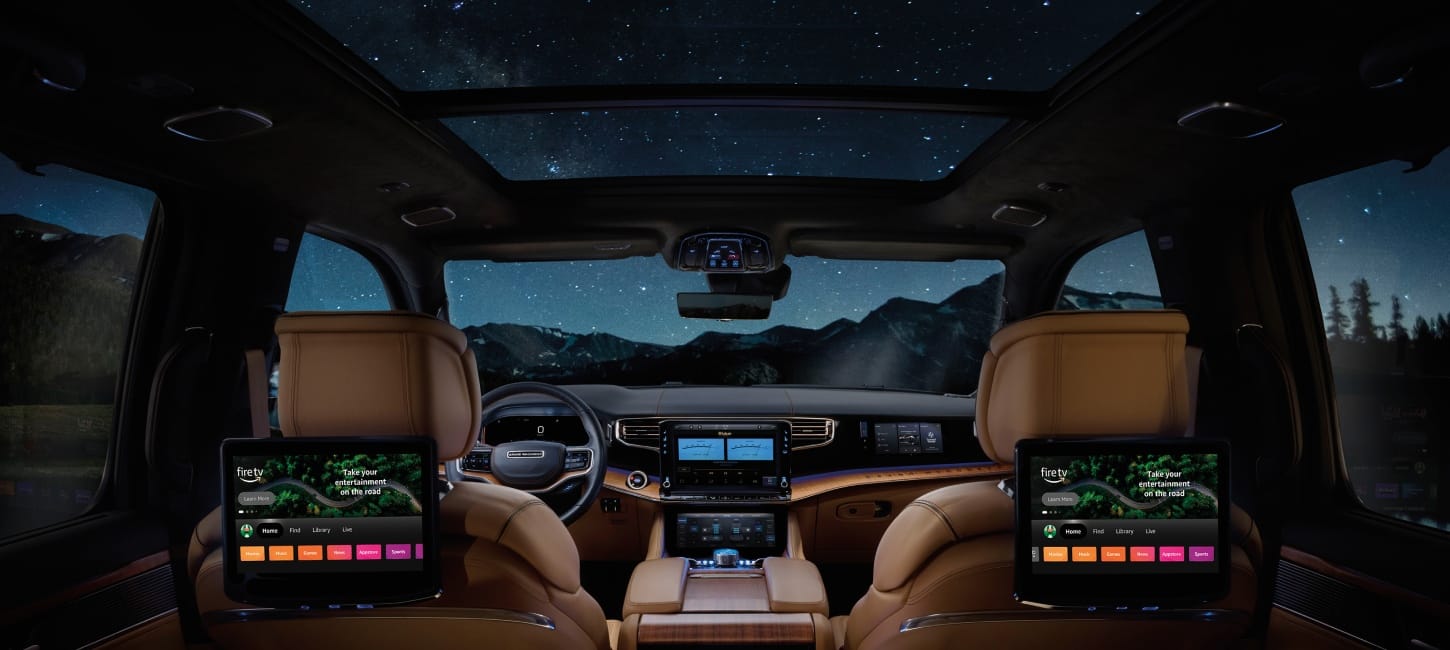 Wide interior view of a Grand Wagoneer Summit Reserve from the rear seat at night, with tan leather-trimmed seats, steering wheel, a panoramic sunroof, and multiple infotainment screens.