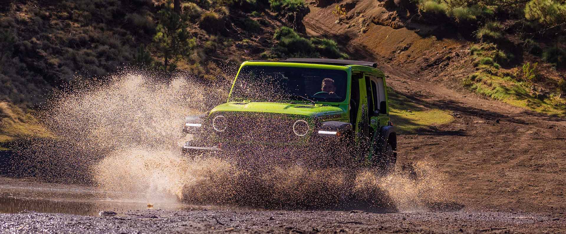 A lime green 2026 Jeep Wrangler Rubicon four-door, entering a stream off-road, kicking up a spray of water as it goes.