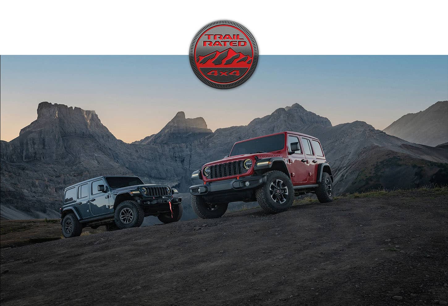 Two four-door 2026 Jeep Wrangler models parked on a clearing off-road. On the left, a gray Wrangler X and on the right, a red Wrangler Rubicon. The Trail Rated badge.