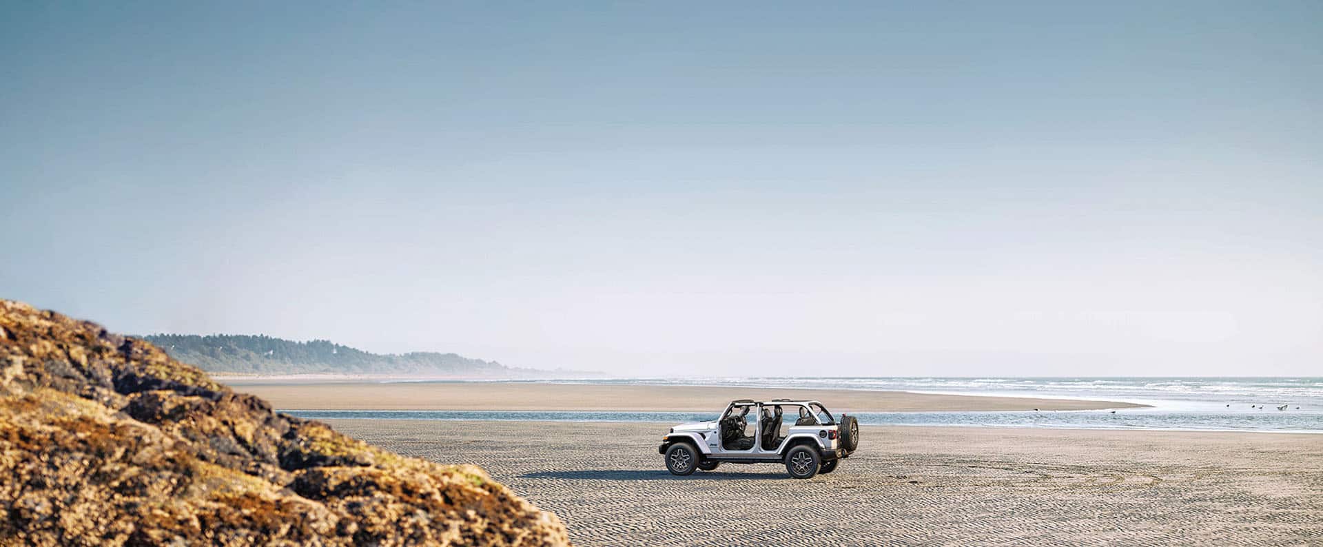 A white 2026 Jeep Wrangler Sahara four-door, with its doors and top removed, parked on a beach.