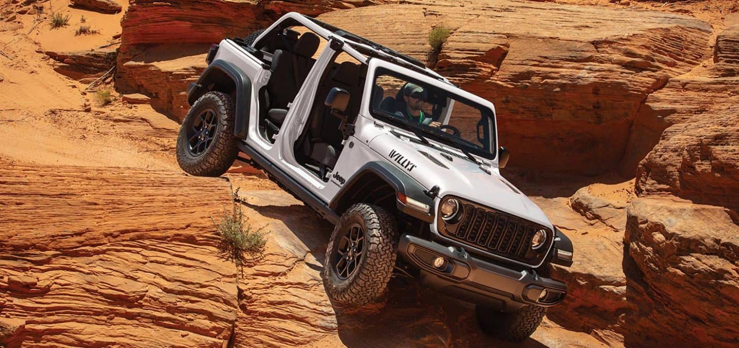 Display A white 2026 Jeep Wrangler Willys four-door, with its doors and top removed, crawling down and over sandstone formations in the desert.