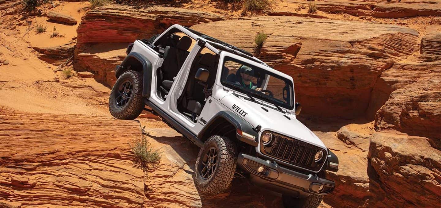 Display A white 2026 Jeep Wrangler Willys four-door, with its doors and top removed, crawling down and over sandstone formations in the desert.