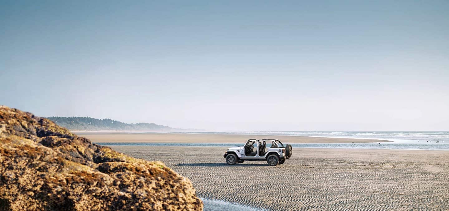 Display A white 2026 Jeep Wrangler Sahara four-door, with its doors and top removed, parked on a beach.