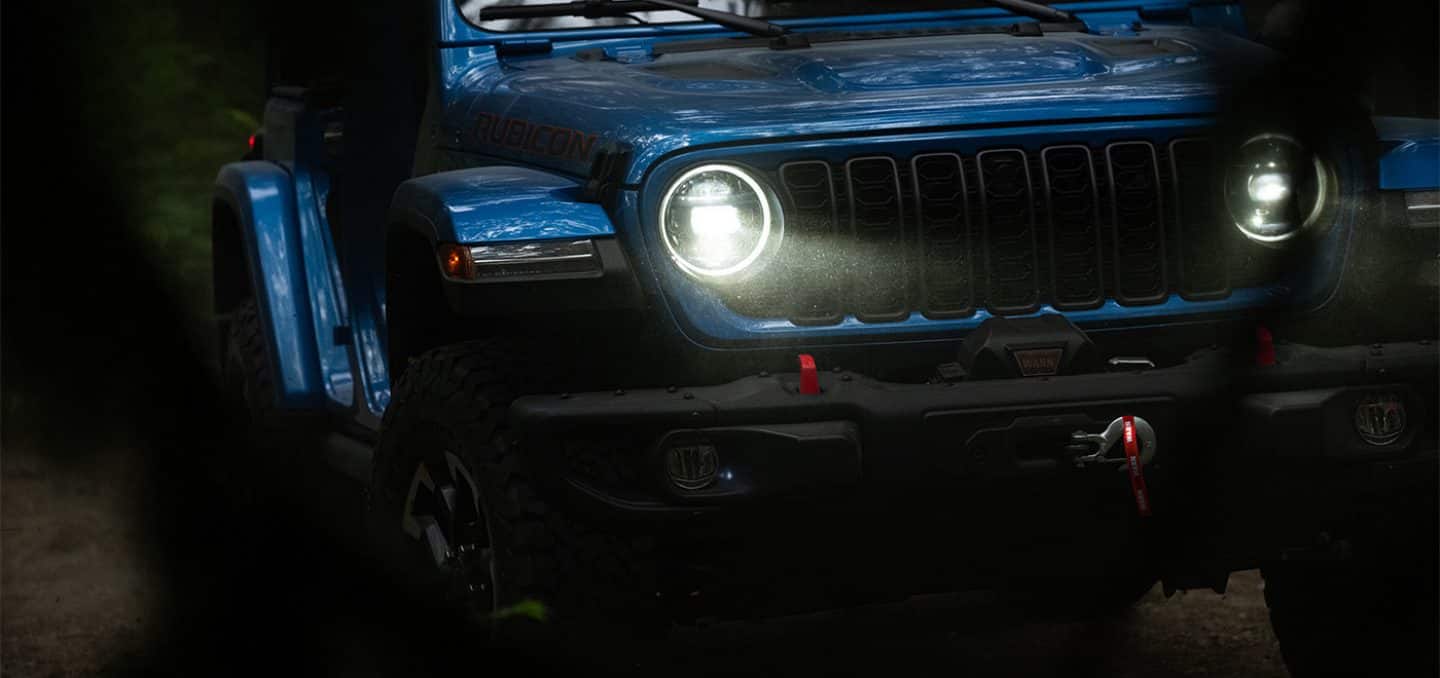 Display The front end of a blue 2026 Jeep Wrangler Rubicon X with its headlamps on.