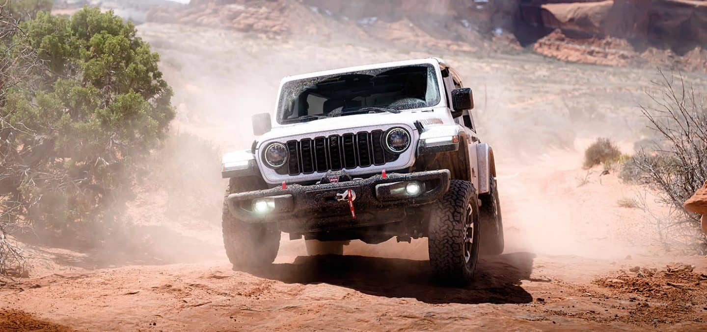 A front angle of a white 2026 Jeep Wrangler Rubicon X traveling on a sandy trail, off-road in the desert.