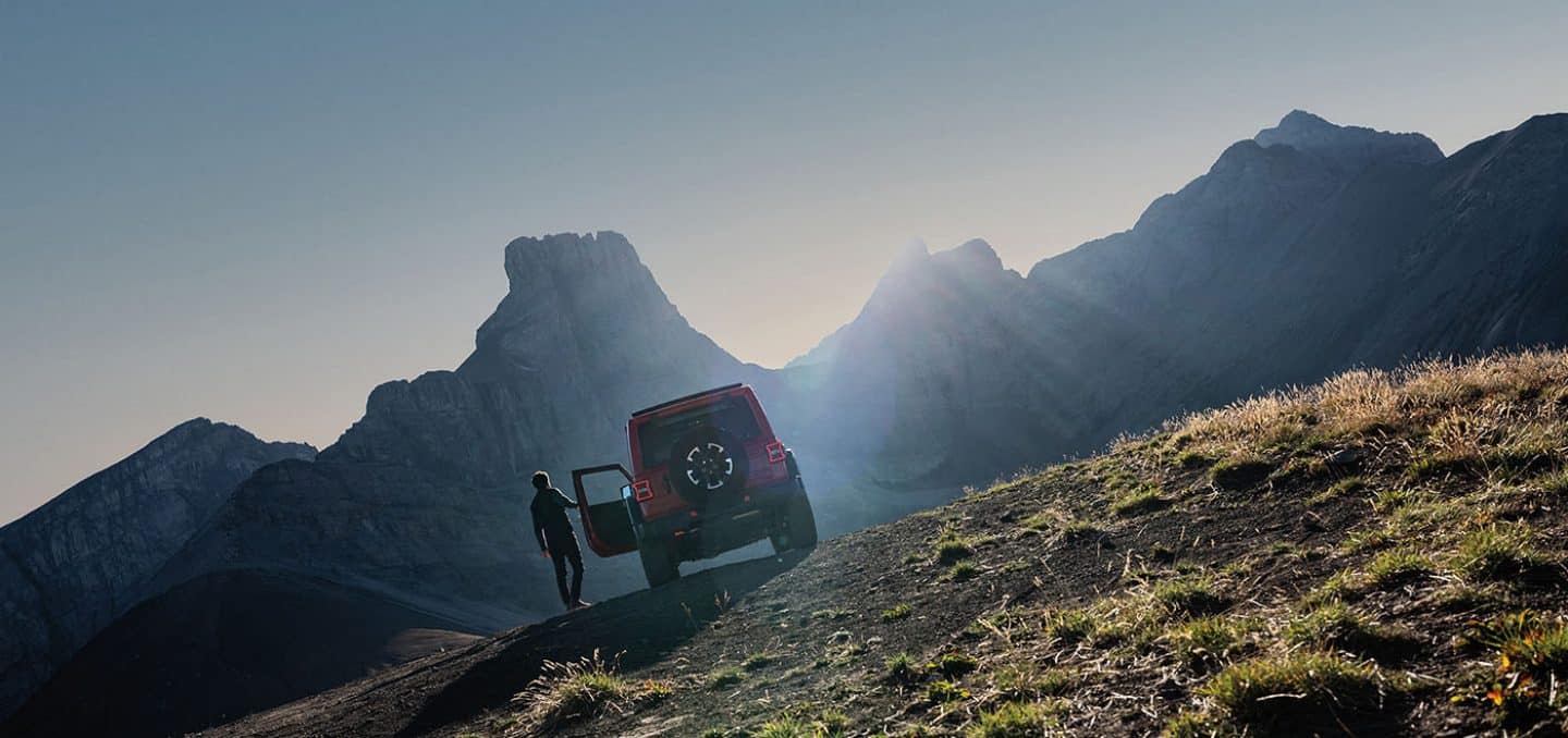 Display A rear angle of a red 2026 Jeep Wrangler Rubicon parked on a clearing off-road in the mountains, with a man opening the driver's door.