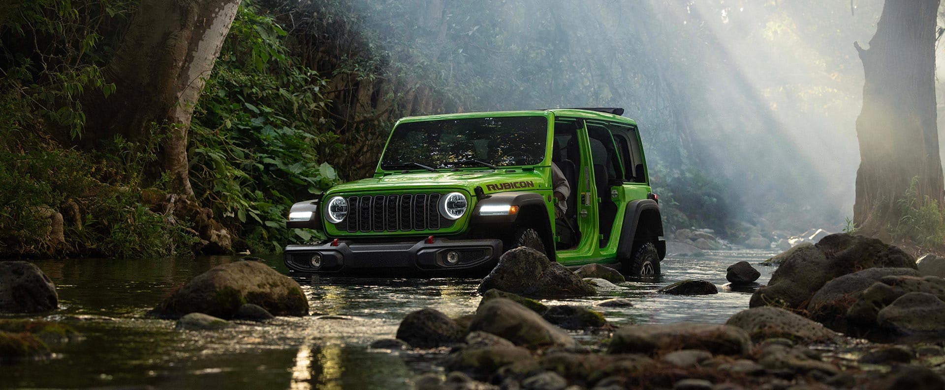 A lime green 2026 Jeep Wrangler Rubicon four-door, with its doors removed, fording through a stream in the woods off-road.