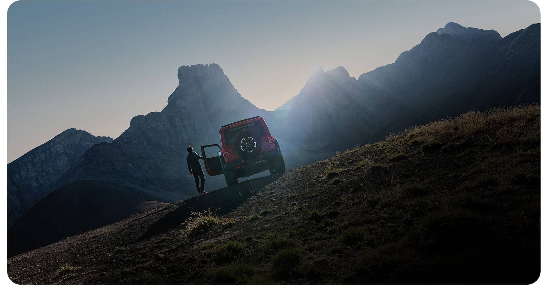 A rear angle of a red 2026 Jeep Wrangler Rubicon parked on a clearing off-road in the mountains, with a man opening the driver's door.