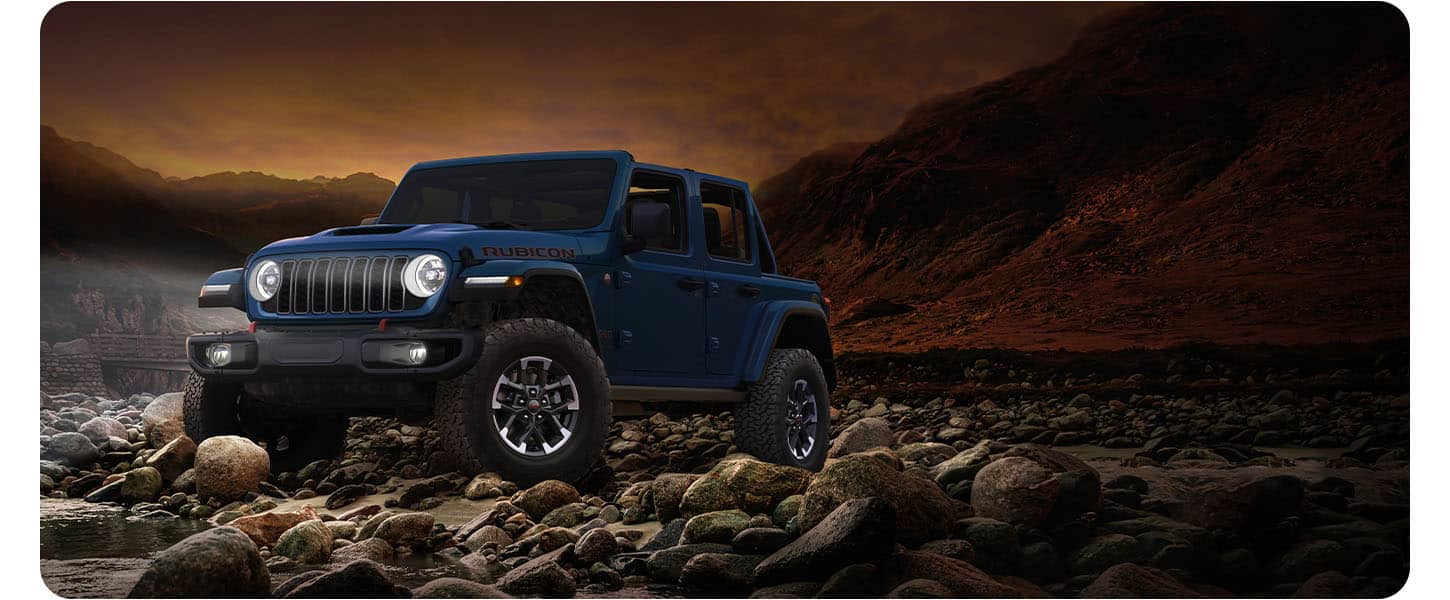 A blue 2026 Jeep Wrangler Rubicon X four-door, with its top removed and its headlamps on, parked on a rocky shoreline at the water's edge at night.