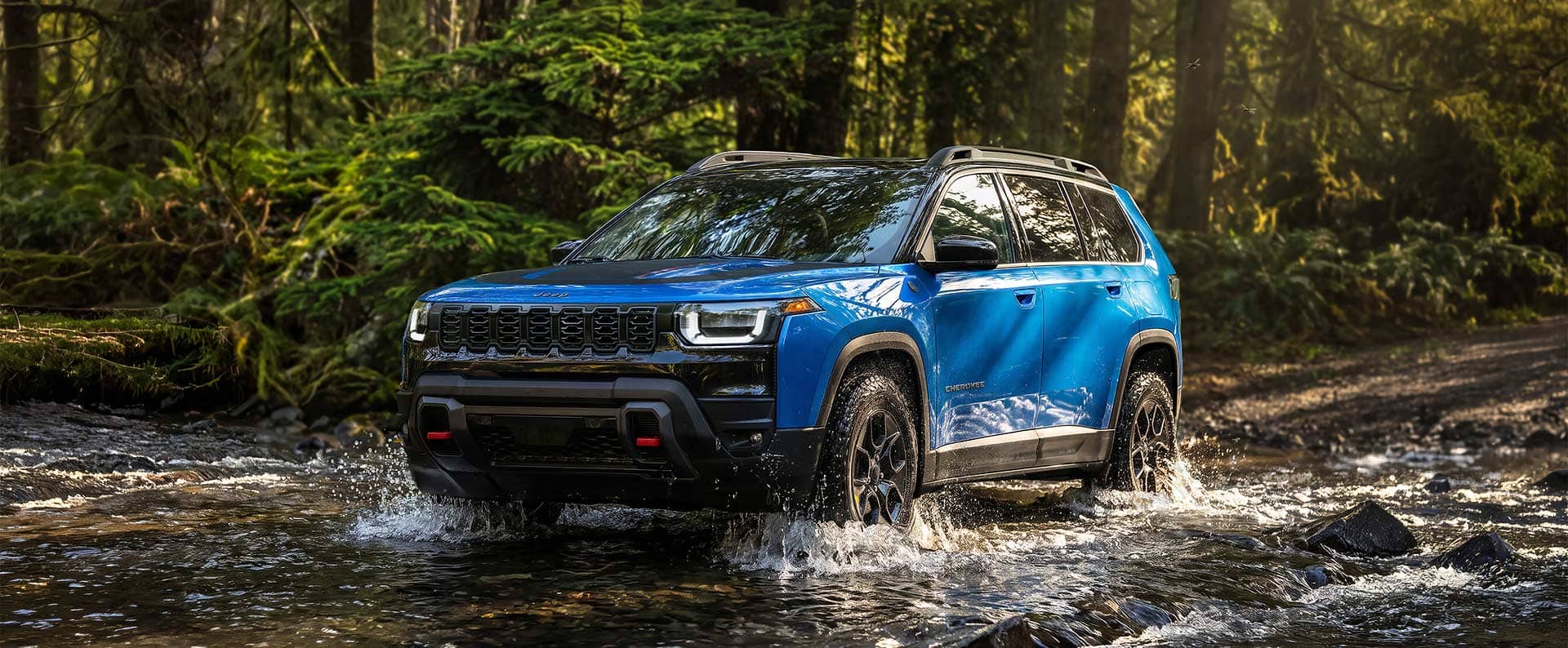 A blue 2027 Jeep Cherokee Trailhawk sprays water as it drives through a creekbed.