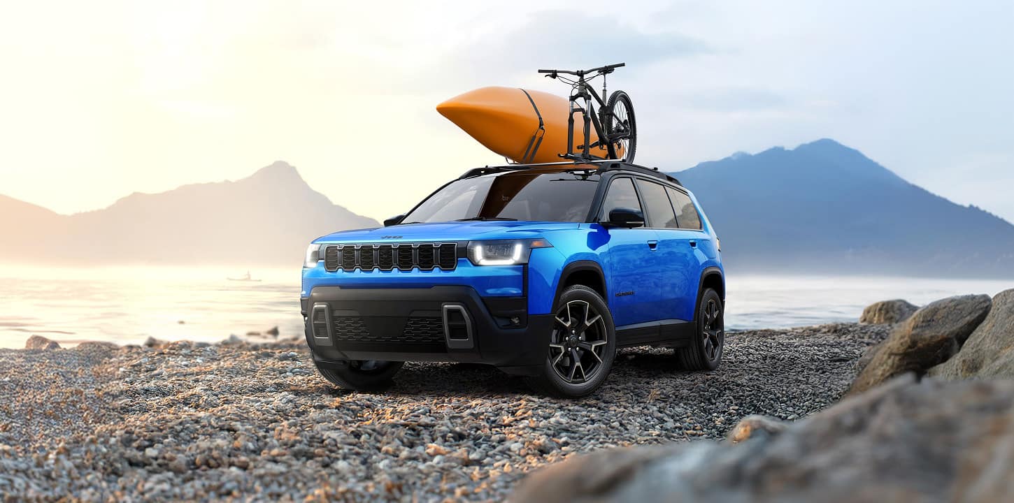 A blue 2027 Jeep Cherokee Overland with a bicycle and an orange kayak on its roof sits parked on a pebble beach.