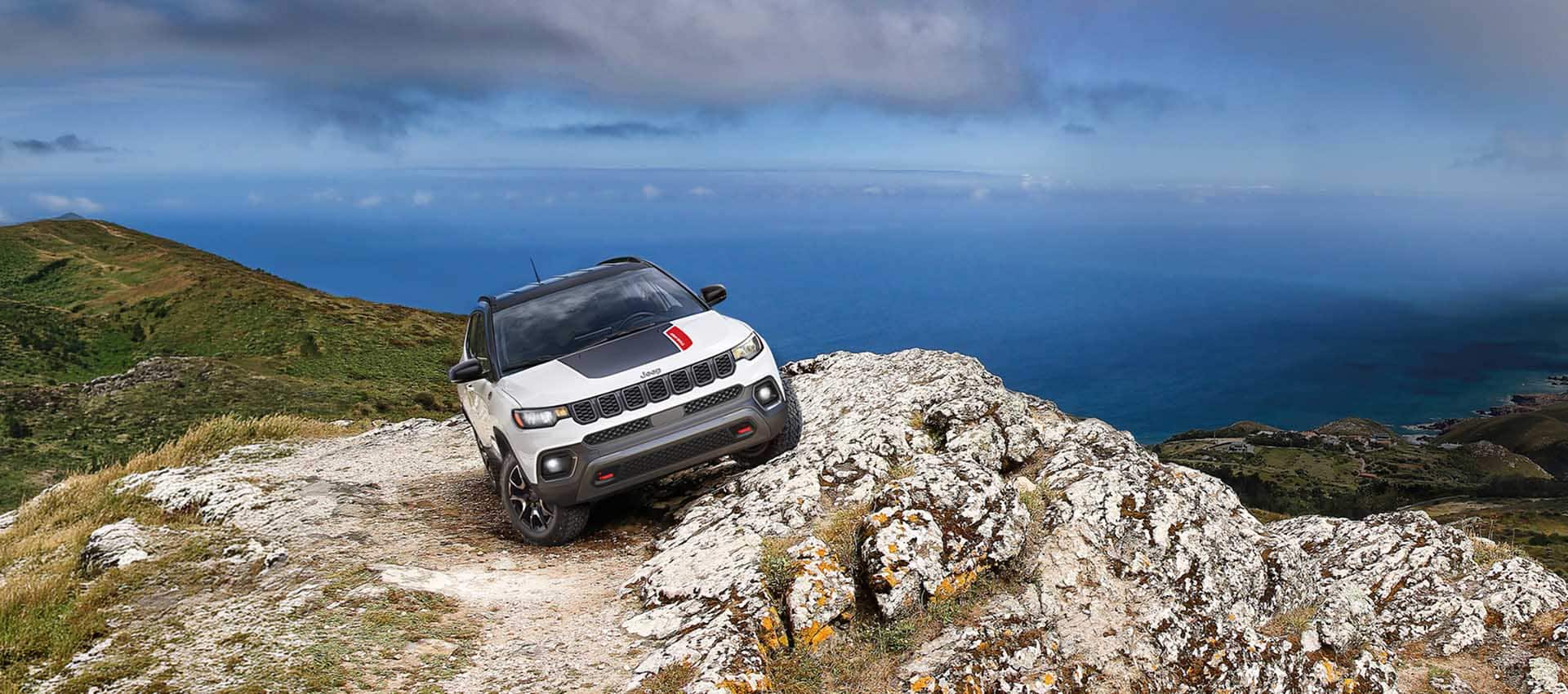 Front view of a white 2027 Jeep Compass Trailhawk on a rocky cliff, overlooking the blue sea during the day.