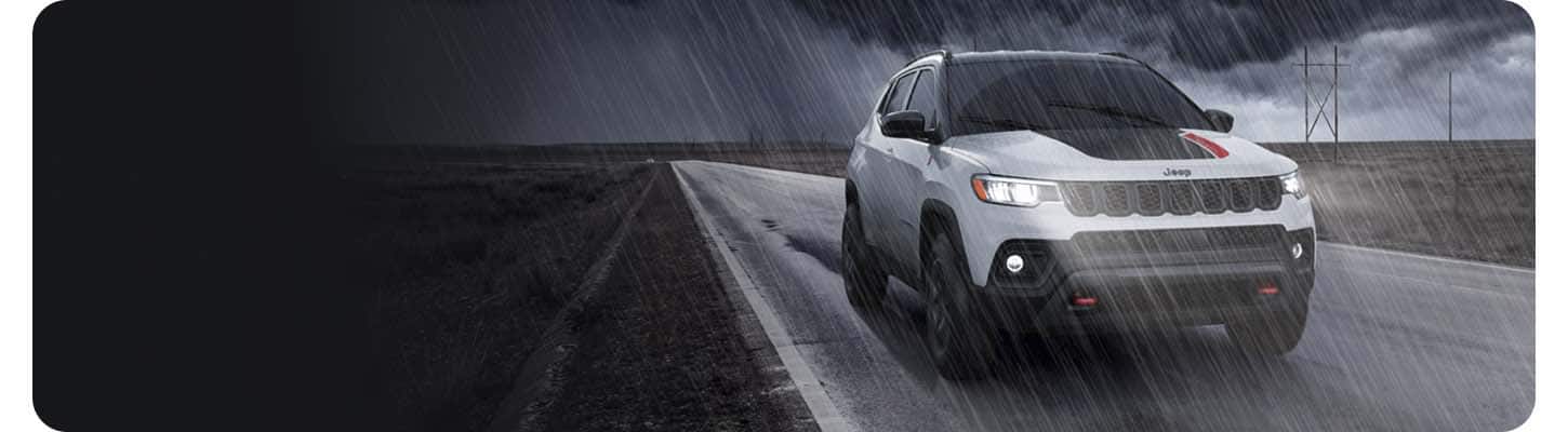 Front three-quarter view of a white 2027 Jeep Compass Trailhawk as it is being driven through the rain on an empty paved rural road.