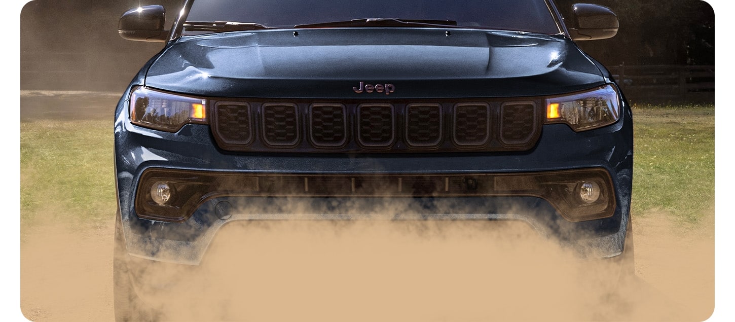 Front view of a dark blue 2027 Jeep Compass Limited Altitude, emphasizing the Jeep grille, surrounded by dust.