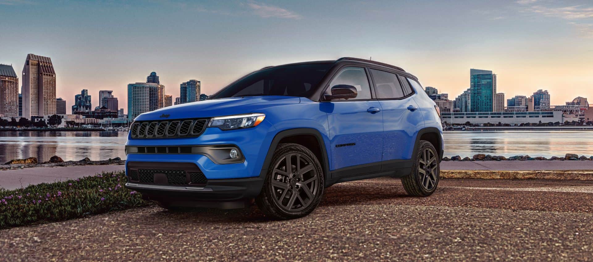 Front three-quarter view of a blue 2027 Jeep Compass Limited Altitude parked on a road in front of a city environment during a sunset.