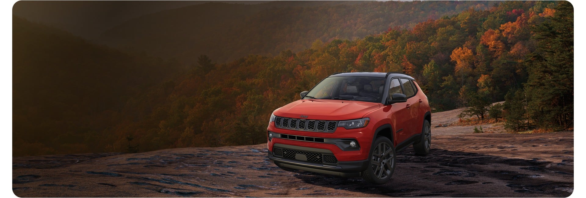 Front three-quarter view of a red 2027 Jeep Compass Limited Altitude parked on a rocky trail surrounded by a hilly tree line during autumn season.