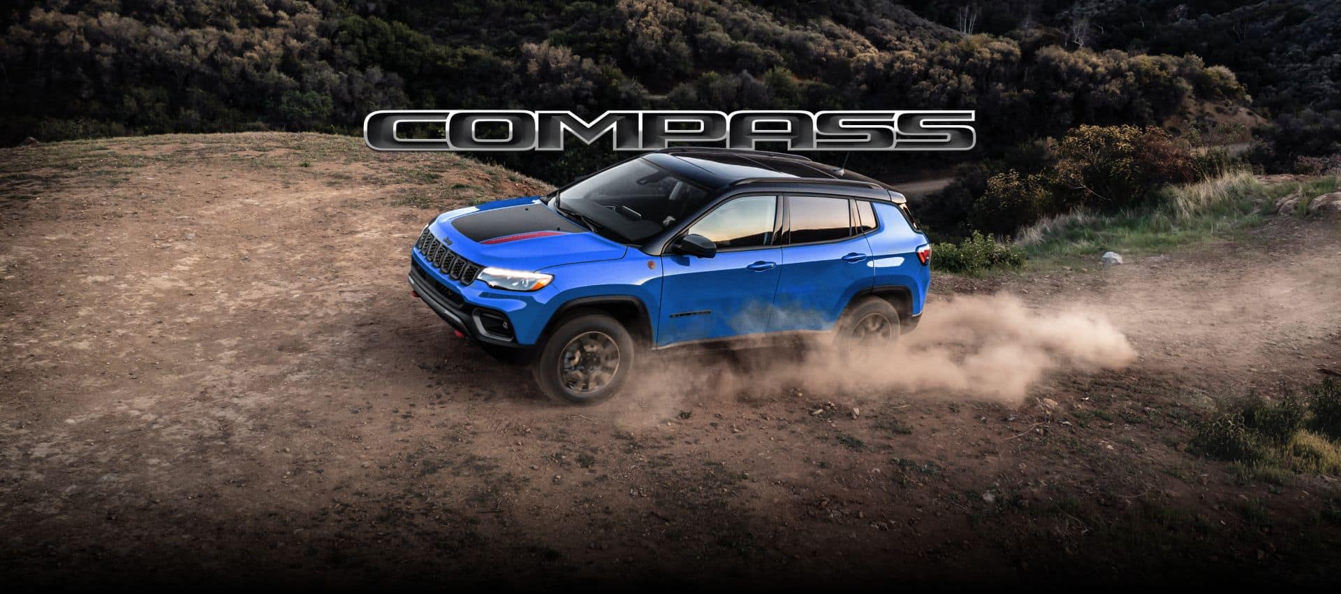 Side view of a blue 2027 Jeep Compass Trailhawk being driven on a dirt trail, kicking up dust.