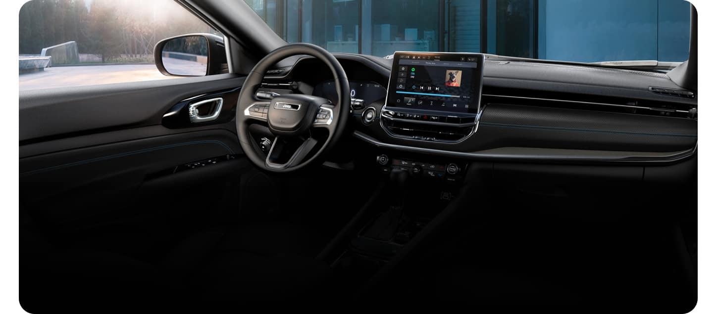 Interior view of a 2027 Jeep Compass showing the driver door, steering wheel, dashboard and center display, with a house visible through the windshield.