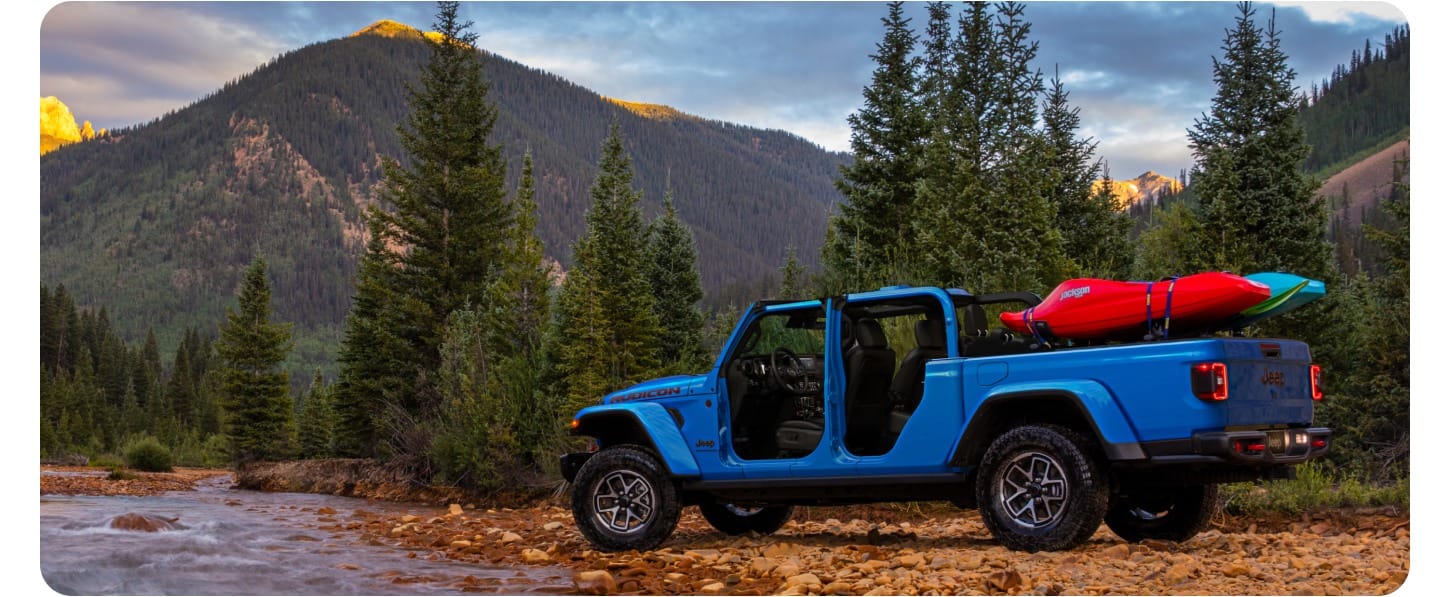 A blue 2027 Jeep Gladiator with the doors off and camping gear on a bed-mounted rack sits parked by the side of a river among wooded mountains.