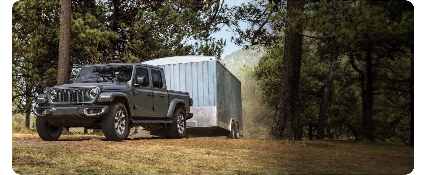 A Gray 2027 Jeep Gladiator Sahara tows a mid-sized trailed on a dirt road in the mountains.