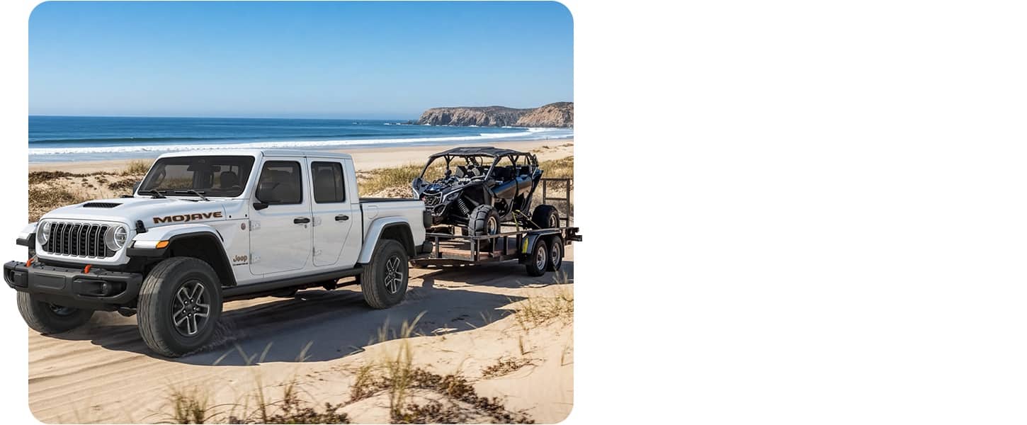 A White 2027 Jeep Gladiator Mojave X tows a trailer with an ATV on it down a sandy around along a shoreline.