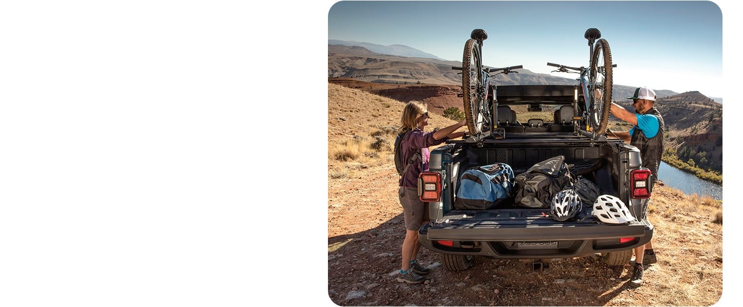 The bed of a 2027 Jeep Gladiator equipped with the Cargo Group with Trail Rail System. Two off-roaders secure their dirt bikes and other gear in the truck bed.