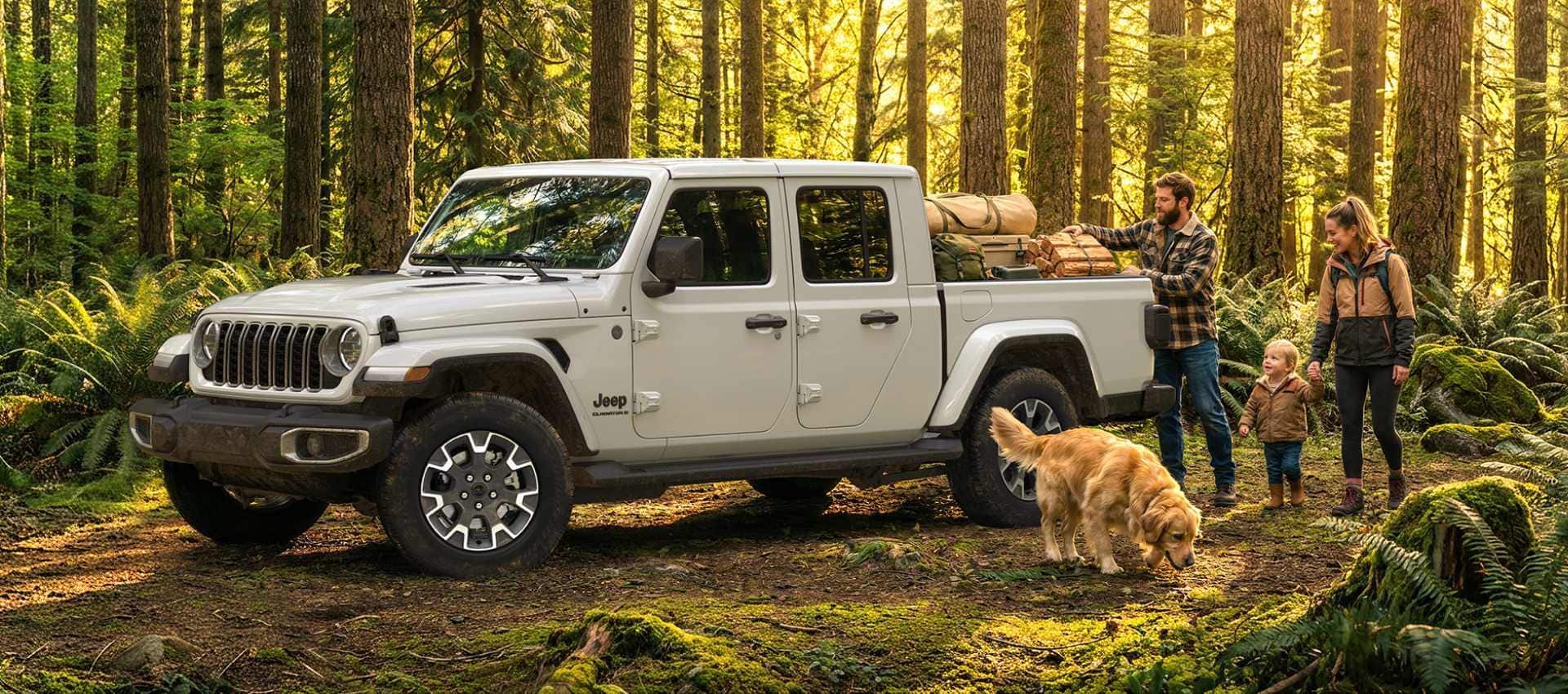 A White 2027 Jeep Gladiator Sahara parked in the forest as a family and their dog begin unloading their gear.
