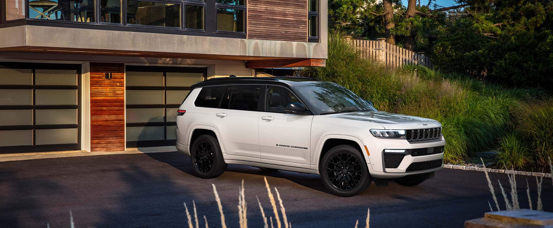 A white and black 2027 Jeep Grand Cherokee L Summit Obsidian parked in the driveway of a contemporary home.