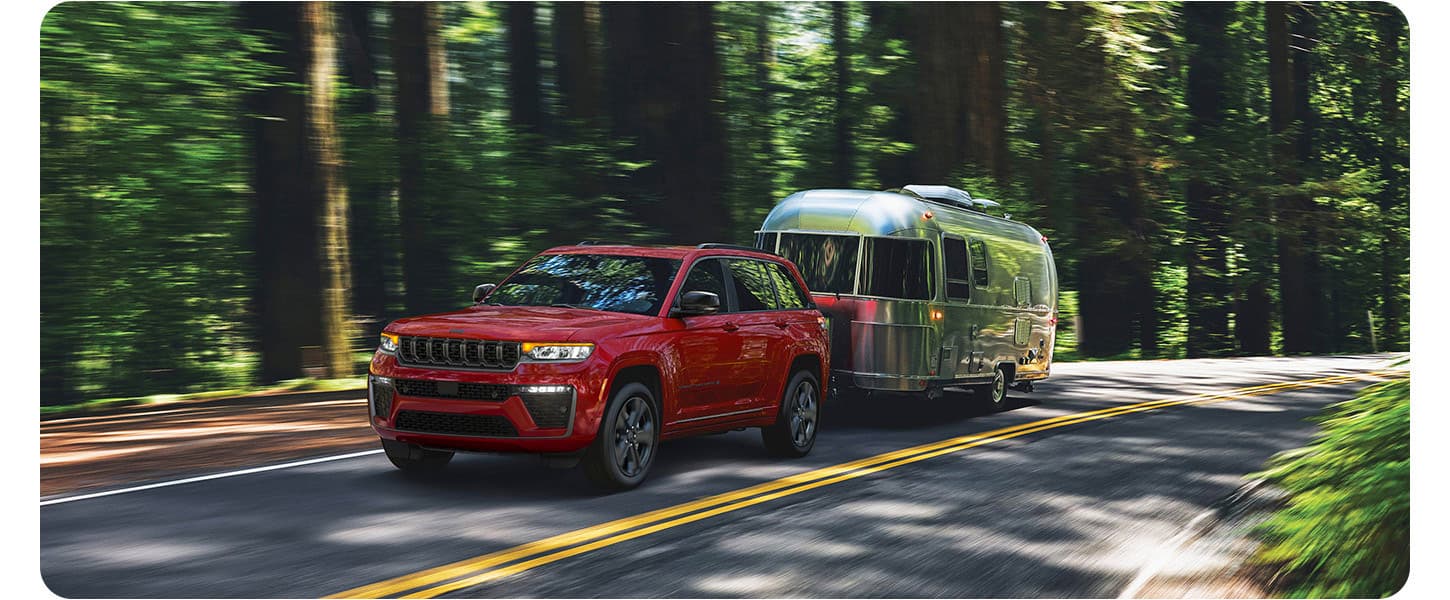 A red 2027 Jeep Grand Cherokee Limited Reserve traveling down a highway in the woods, towing a travel trailer.