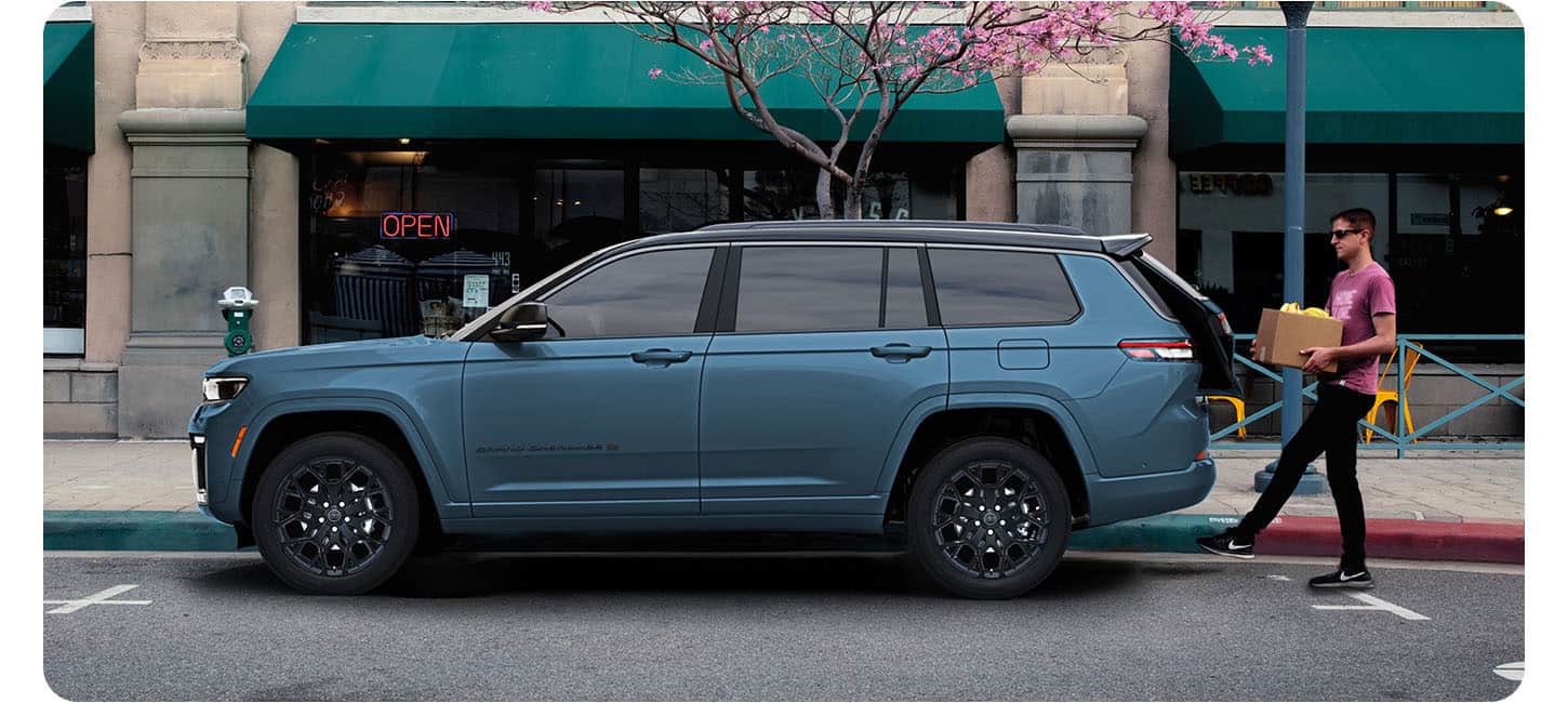 A driver-side profile of a blue 2027 Jeep Grand Cherokee Summit Obsidian parked on a city street in an area of shops as a man with a box in his hands, swings his foot under the liftgate, opening it hands-free.