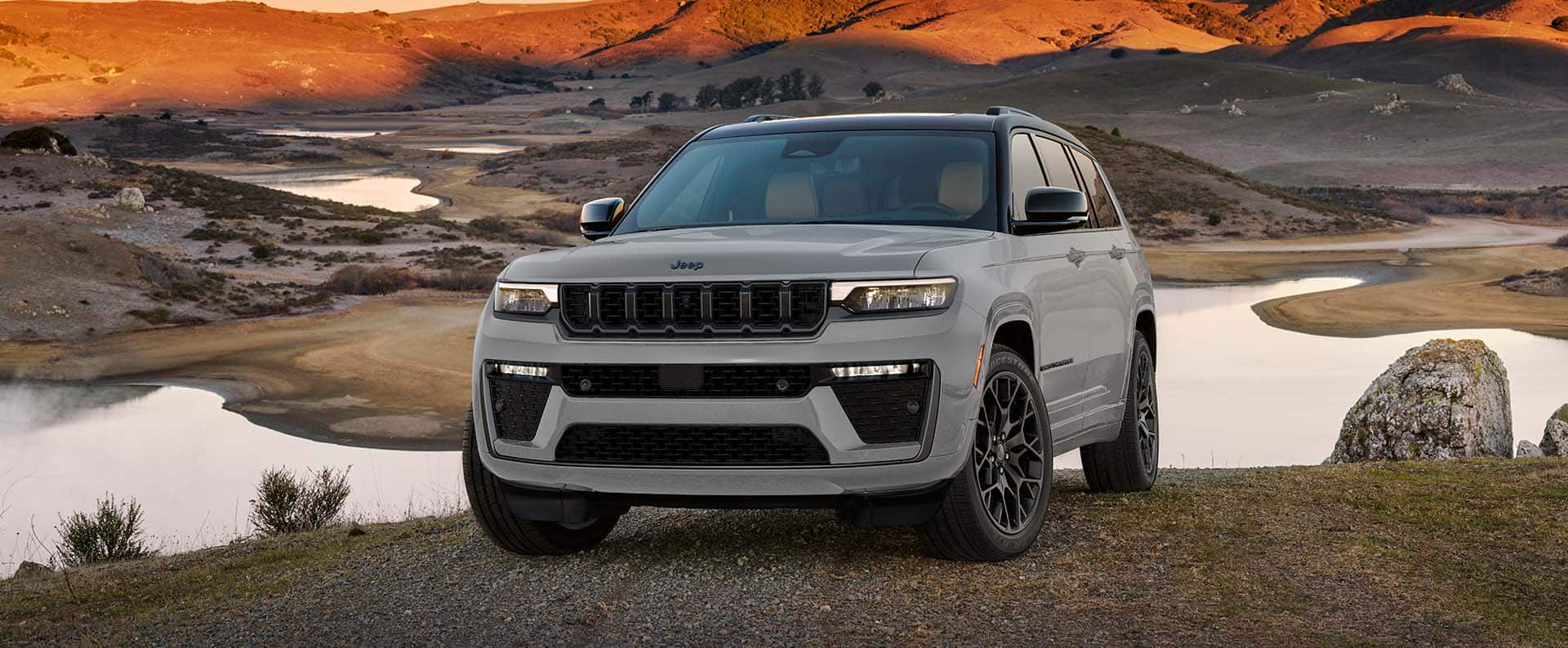 A gray 2027 Jeep Grand Cherokee Summit Obsidian parked on a hill overlooking a lake during sunset.