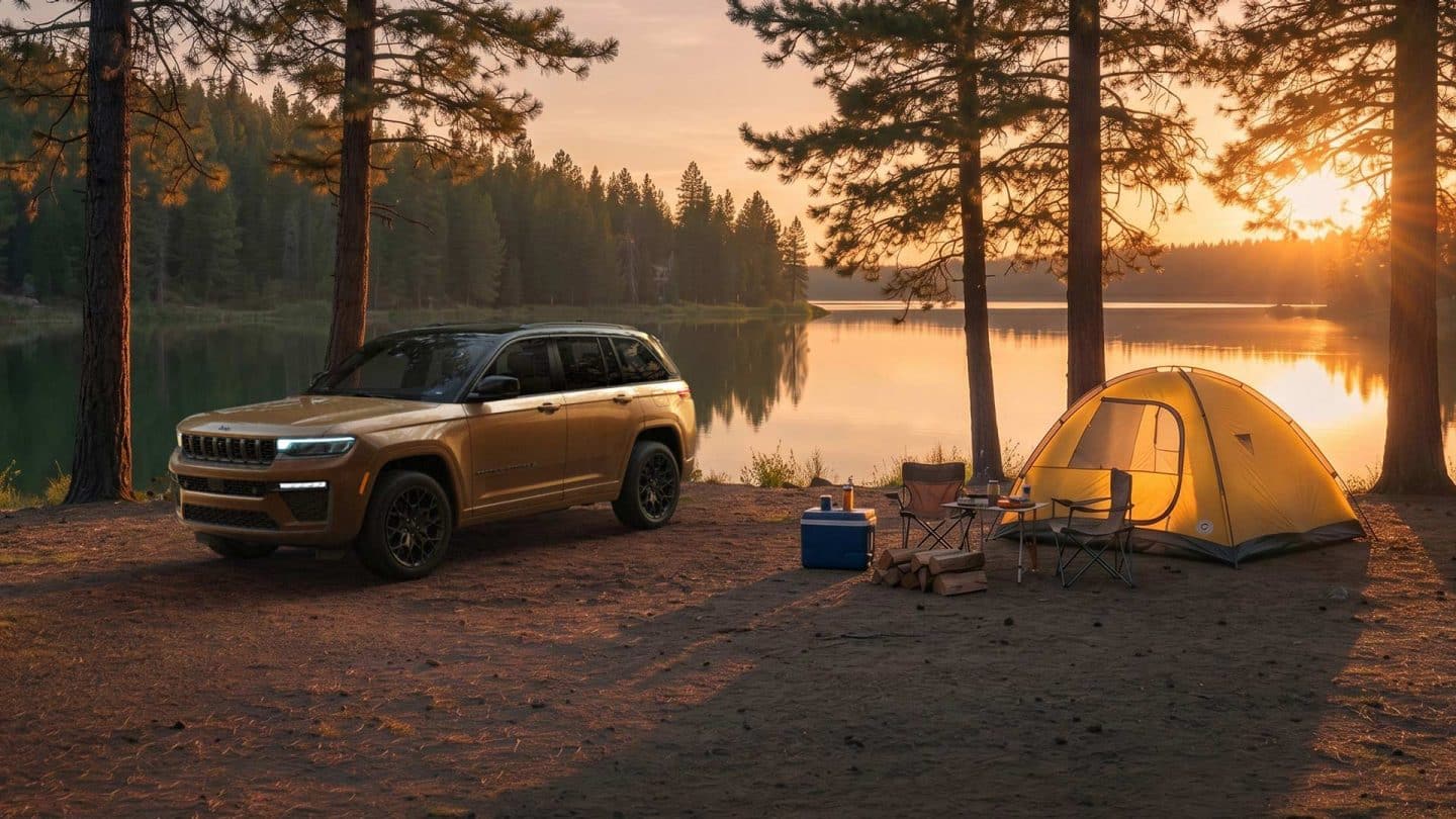 A copper and black 2027 Jeep Grand Cherokee Summit Obsidian parked at a campsite beside a lake at sunset.