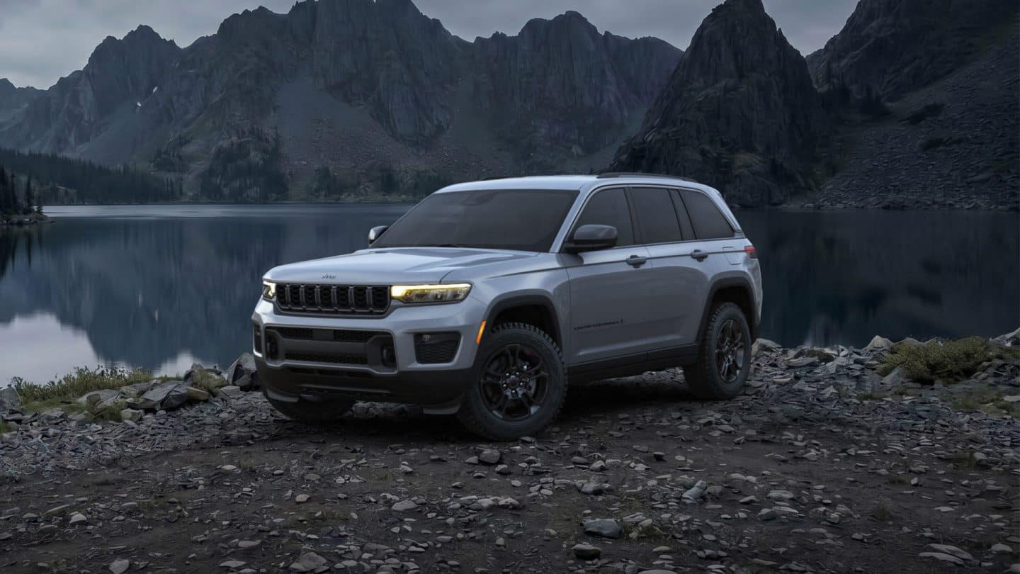 A gray 2027 Jeep Grand Cherokee Upland parked on a rocky beach beside a lake at night.