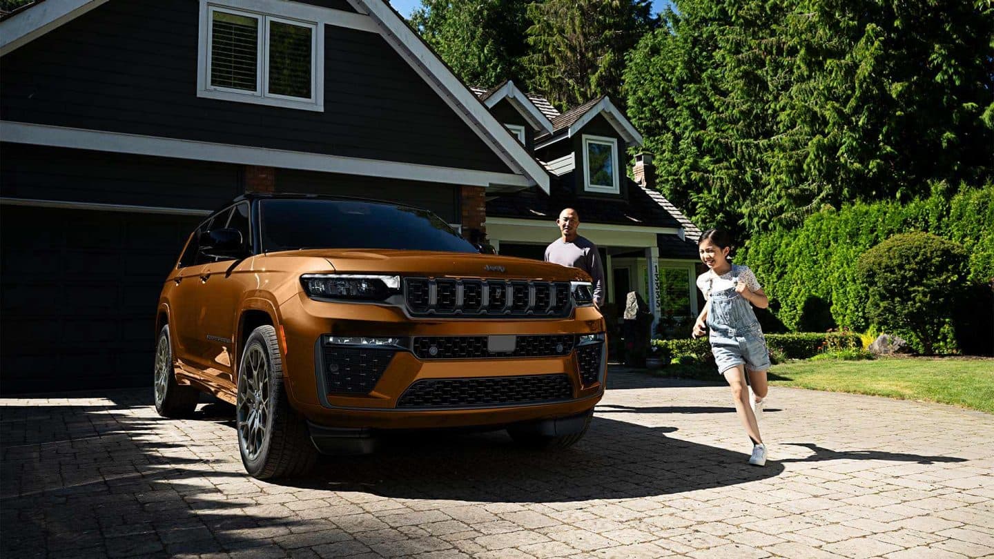 A copper and black 2027 Jeep Grand Cherokee Summit Obsidian parked in the driveway of a residential home with a man and his daughter nearby.