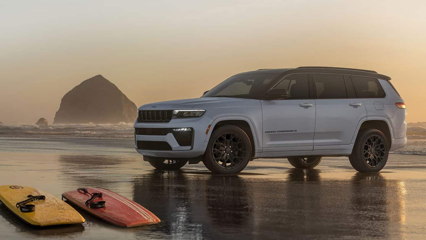 A silver and black 2027 Jeep Grand Cherokee Summit Obsidian parked on a foggy beach at sunrise with two boogie boards nearby.