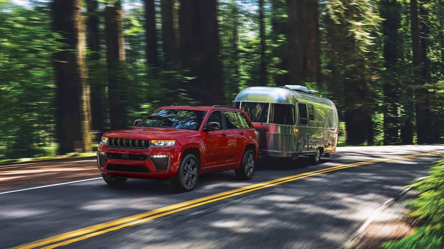 A red 2027 Jeep Grand Cherokee Limited Reserve traveling down a highway in the woods, towing a travel trailer.