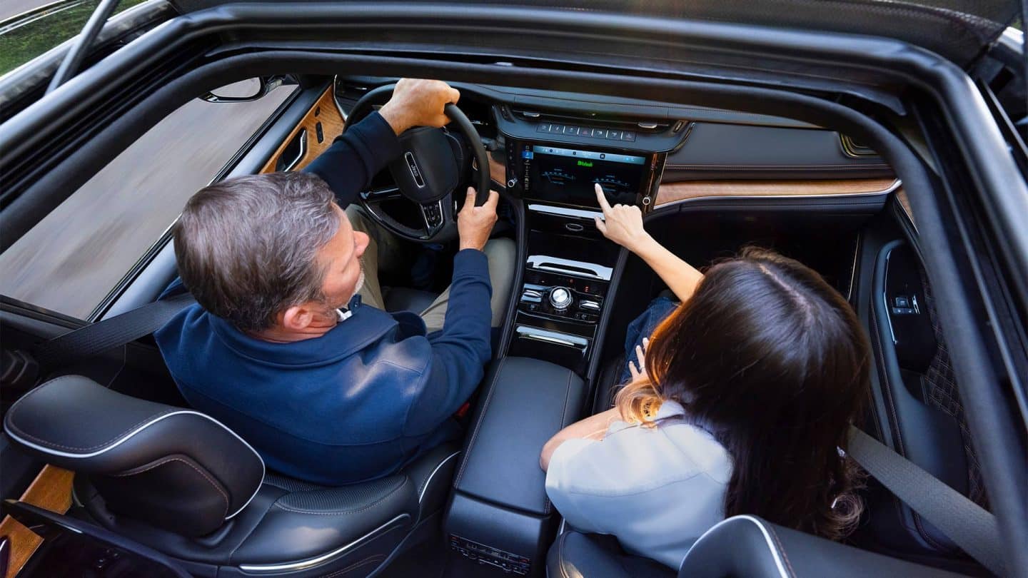 The front seat area of the 2027 Jeep Grand Cherokee Summit, with a man in the driver seat and a woman in the passenger seat. Shot from outside the open sunroof.