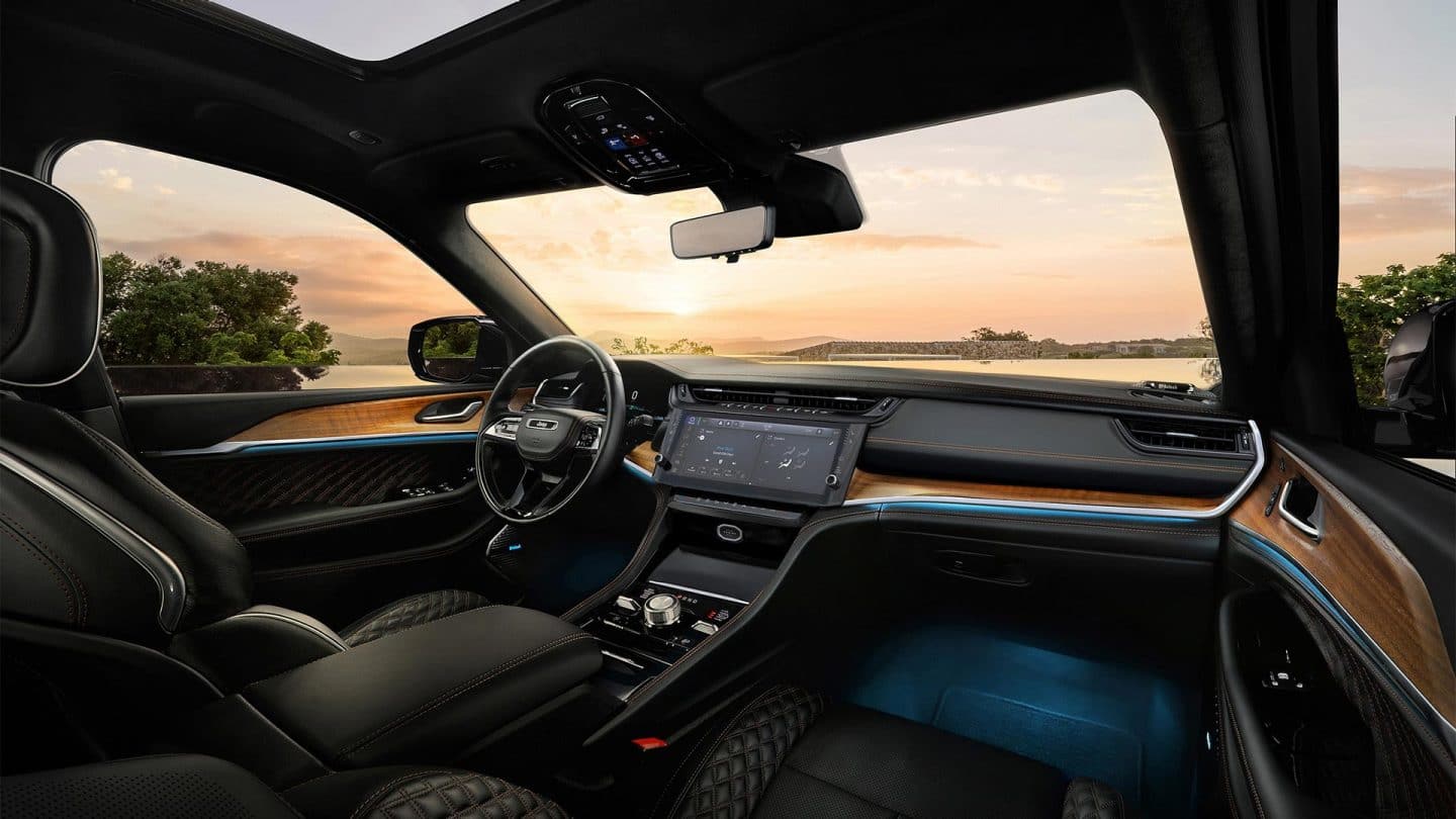 The front seat area of the 2027 Jeep Grand Cherokee Summit, focusing on the driver seat, steering wheel, Digital Cluster, Uconnect touchscreen, center stack controls center console and the view of the sunset through the windshield.